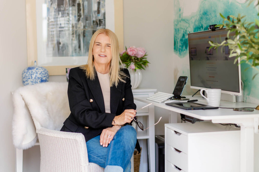 a photo of Yvette Craig sat at her desk in jeans and a jacket holding her glasses